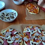 Dwie pizze na surowym cieście z salami, szynką i pieczarkami na papierze do pieczenia. Dzieci dodają składniki w tle. Kolorystyka ciepła, dominują brązy i beże.