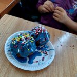 Dwa pączki z niebieskim lukrem i kolorową posypką leżą na papierowym talerzu. Tło to drewniany stół. Za nimi dziecko w fioletowej bluzie.