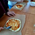 Dzieci przygotowują mini pizze na brązowym stole. Jasne ciasto z sosem pomidorowym leży na papierze do pieczenia. Obok jest miska z szynką.