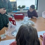 received_6508347047454781 Osoba w zielonej sukience używa czerwonej prasy ręcznej. Dzieci siedzą przy stole z materiałami plastycznymi. W tle jasne okno i rośliny.