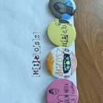 Cztery kolorowe przypinki na białej kopercie z napisem MIŁOŚĆ. Tło stanowi drewniany blat. Kolory: niebieski, żółty, pomarańczowy, różowy.