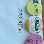 received_6521440379502741 Cztery kolorowe przypinki leżą na białej kopercie. Mają dziecięce rysunki i napisy, m.in. MIŁOSZ. Koperta jest lekko zmięta.