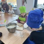 received_7358083625765231 Dzieci w kolorowych czapkach kucharskich i fartuchach siedzą przy stole. Na stole miska z ciastem; tło stanowi jasne wnętrze z oknem. Dzieci wyglądają na skupione, są w trakcie gotowania.