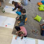 received_7849872179667281 Na zdjęciu czworo dzieci rysuje na dużych kartkach papieru w kolorowym pokoju. Tło stanowi szary dywan oraz jasne drewniane podłogi. Obok dzieci leżą kolorowe poduszki i pudełko z kredkami.