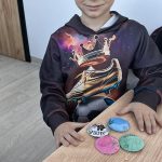 received_7897043208205041 Chłopiec w kolorowej bluzie z butem i koroną siedzi przy stole. Przed nim leżą kolorowe, własnoręcznie ozdobione medaliony. Tło jest jasne, ściany białe, podłoga drewniana.