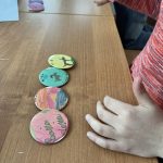 received_8117202218786131 Na drewnianym stole leżą cztery kolorowe okrągłe znaczki. Dziecko w czerwonej bluzce trzyma jeden w dłoni. Na znaczkach są różne kolorowe wzory.