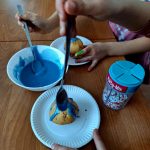 received_8542723307519071 Ciemnobrązowy stół z dziećmi oblewającymi babeczki niebieskim lukrem i posypką. Na talerzach miseczki. Obok pojemnik z posypkami.