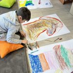 received_8607324301718461 Chłopiec rysuje kredkami na dużych kartkach, siedząc na dywanie obok pomarańczowej poduszki. Rysunki są kolorowe, z dominacją czerwieni, niebieskiego i zieleni. Tło to jasna podłoga z drewnianym regałem po prawe