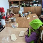 Grupa dzieci w kolorowych czapkach kucharza siedzi przy stole. Na drewnianym blacie są drewniane deski, mąka i miska. W tle stoją szafki z przyborami kuchennymi.