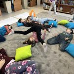 Na dywanie w bibliotece leży grupa dzieci w kolorowych ubraniach. Dookoła są kolorowe poduszki: niebieskie, zielone, pomarańczowe. W tle regały z książkami i zielone rośliny.