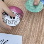 received_907855868344498 Małe dziecko trzyma w rękach dwa kolorowe jojo na drewnianym stole. Jeden jest zielony, drugi różowo-biały z literami. Obok leży papier z napisem.