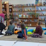 Dzieci słuchają siedząc na poduszkach w bibliotece pełnej książek. Nauczycielka w kolorowym ubraniu pochyla się nad nimi. Jasne ściany i wygodne fotele dodają przytulności.