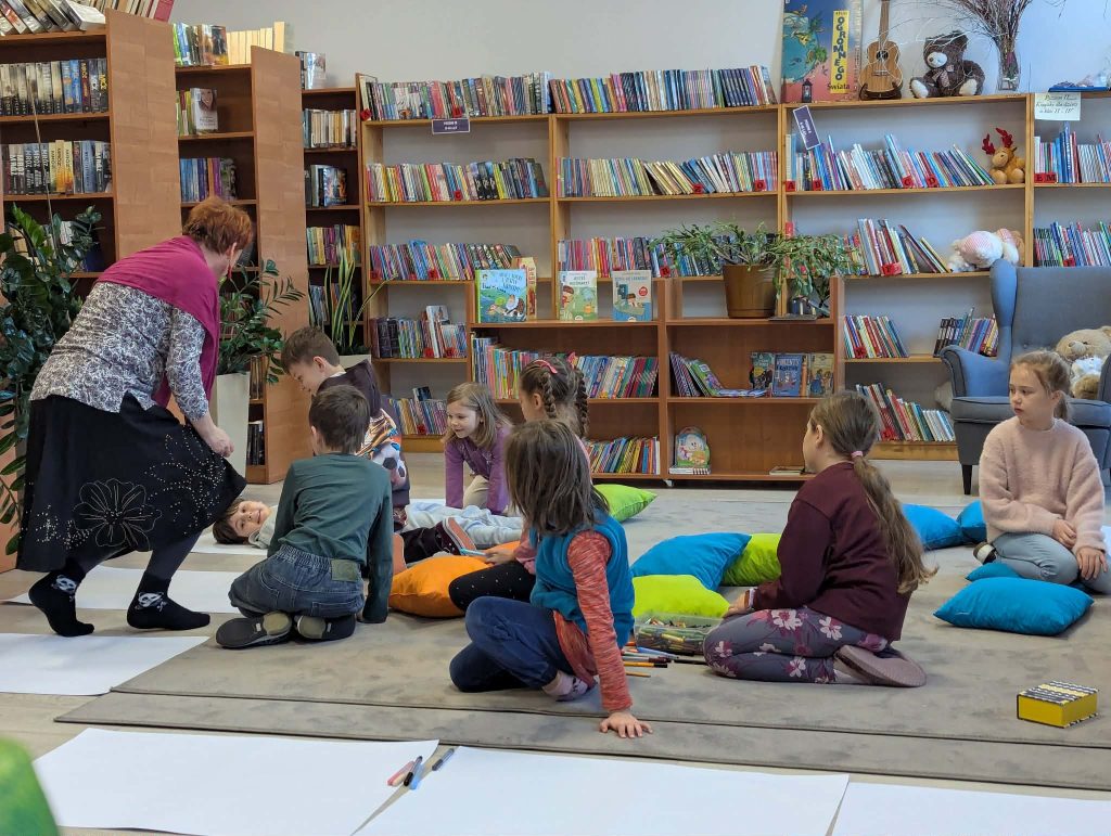 Grupa dzieci siedzi na poduszkach w kolorowej bibliotece. Nauczycielka w różowym szaliku pomaga leżącemu dziecku. W tle regały z książkami i dekoracje.