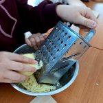 Dziecko trze ser na srebrnej tarce nad metalową miską. Dłoń w bordowej bluzie, tło rozmyte, jasne powierzchnie.