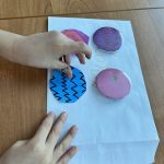 received_9279860465636681 Dziecko układa kolorowe papierowe koła na białej kartce. Tło to drewniany stół. Koła są w odcieniach niebieskiego i różowego.