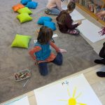 Grupa dzieci rysuje na podłodze pokrytej szarym dywanem. Obok leżą kolorowe poduszki: niebieskie, zielone i pomarańczowe. W tle widoczny regał z książkami.