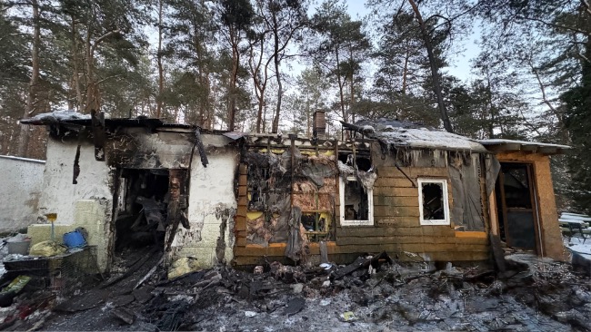 Spalony dom w lesie, otoczony drzewami. Ściany są czarne od ognia, a dach częściowo zawalił się. Ziemia jest pokryta popiołem.