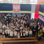 Grupa ludzi w sali, dzieci i dorośli w białych koszulach, przed tłem z polską flagą i napisem o bitwie pod Dobrem.