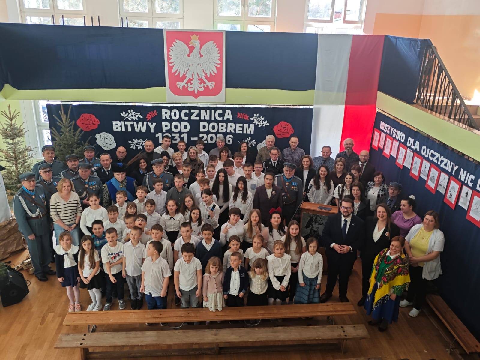 Grupa ludzi w sali, dzieci i dorośli w białych koszulach, przed tłem z polską flagą i napisem o bitwie pod Dobrem.