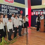 637872885_931741265868062_3371352594162972512_n Grupa dzieci w białych koszulach śpiewa na tle ciemnej kurtyny z napisem o rocznicy bitwy. Widać flagę Polski i rysunki na ścianie. Drewniana podłoga, mikrofony i dekoracje w odcieniach czerwieni i bieli