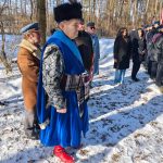 637884025_931740779201444_6584766794623860413_n Mężczyzna w niebieskim stroju z piórami stoi na śniegu. Obok inni w zimowych ubraniach. W tle drzewa w zimowym krajobrazie.