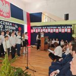 637885886_931741379201384_7867449278681857919_n Grupa dzieci w białych koszulach występuje na scenie ozdobionej flagą Polski i herbem. Tło w kolorach biało-czerwonych. Na widowni siedzą dorośli, a obok sceny znajduje się podium.