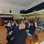 637917217_931741325868056_3094540645596980059_n Na zdjęciu widać szkolną uroczystość z uczestnikami siedzącymi na ławkach. Tło dekorowane jest granatową tkaniną z białym napisem. Sala ma jasne ściany i drewnianą podłogę.