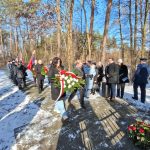 638289711_931740265868162_9022511823364457966_n Grupa osób składa kwiaty na ośnieżonym chodniku w lesie. Dominują kolory zielony, biały i czerwony. Na zdjęciu są także flagi i uczestnicy w ciemnych płaszczach.