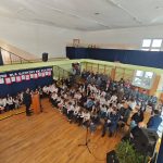 638720063_931741555868033_8995524905320580969_n Sala gimnastyczna z wystrojem patriotycznym. Uczniowie w białych koszulach siedzą na krzesłach. Na scenie mówca przy pulpitu.