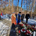 639443629_931740712534784_5377166371025875265_n Grupa ludzi w zimowych płaszczach stoi przy pomniku w lesie, na tle śnieżnym. Kwiaty i znicze są ułożone z przodu. Tło stanowią wysokie drzewa i błękitne niebo.