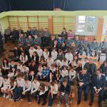 639673017_931742085867980_4044350560613491377_n Zdjęcie ukazuje zgromadzenie ludzi w sali gimnastycznej. Dzieci i dorośli siedzą na ławkach, z przodu kilka osób w mundurach. Tło w jasnych kolorach z oknami i drabinkami gimnastycznymi.