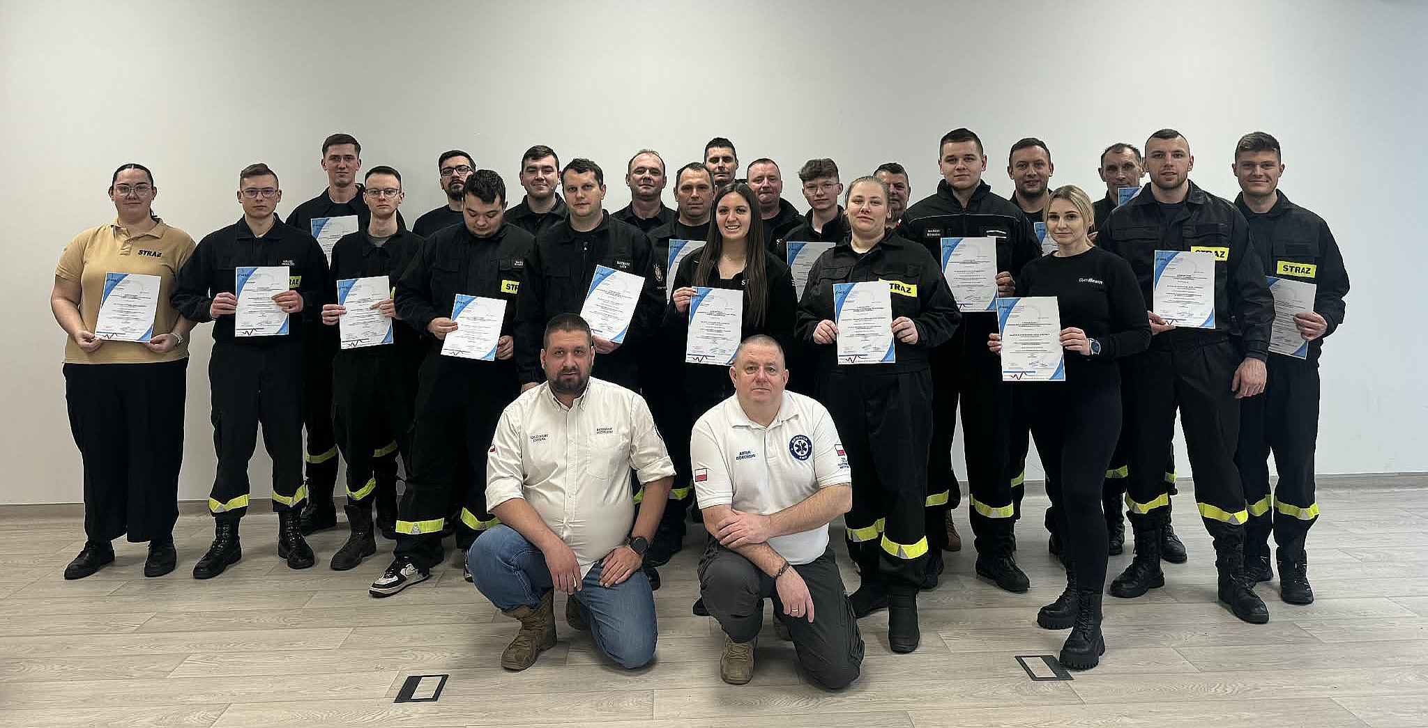 Grupa osób w czarnych mundurach trzyma certyfikaty. Stoją na jasnym tle. W centrum dwóch mężczyzn klęczy.