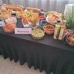 20260315_162236 Stół z czarnym obrusem pełen przekąsek: pieczywa, nachosów, warzyw, sosów oraz oliwy. Obok zioła w doniczkach. Tło w jasnych kolorach.