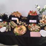 20260315_162308 Na czarnym obrusie znajdują się półmiski z różnorodnymi przekąskami. Kolorowe dania są udekorowane warzywami i ziołami. W tle białe ściany oraz ozdobne świece i kwiaty.