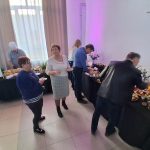 20260315_165428 Na zdjęciu widzimy ludzi przy bufecie, wybierających jedzenie. W tle zasłonięte okno i ściana w jasnych kolorach. Stoły pokryte są czarnymi obrusami, a dania są różnorodne.