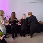 20260315_165551 Cztery osoby stoją przy stole szwedzkim z ciemnym obrusem. Na stole są kwiaty, przekąski i świeczka. Tło jasne, ambient różowy.