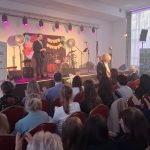 20260315_171720 Na scenie jasne oświetlenie, stojący mężczyzna w garniturze, dekoracje z kwiatów. Publiczność siedzi na czerwonych krzesłach. Obok sceny biały baner z napisem „Mazowsze”.