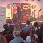 20260315_171727 Mężczyzna w garniturze przemawia na scenie przy mikrofonie. Za nim widoczny jest zespół perkusyjny i jasne dekoracje. Na pierwszym planie siedzi publiczność.