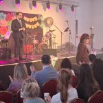 20260315_171744 Osoba na scenie przemawia do publiczności. Tło zdobią kolorowe dekoracje i instrumenty muzyczne. W pomieszczeniu panuje przytulna atmosfera.