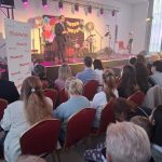 20260315_171946 Mężczyzna na scenie w ciemnym garniturze przemawia do publiczności. Scena udekorowana kolorowymi kwiatami. W tle widoczne instrumenty muzyczne, a publiczność siedzi na czerwonych krzesłach.