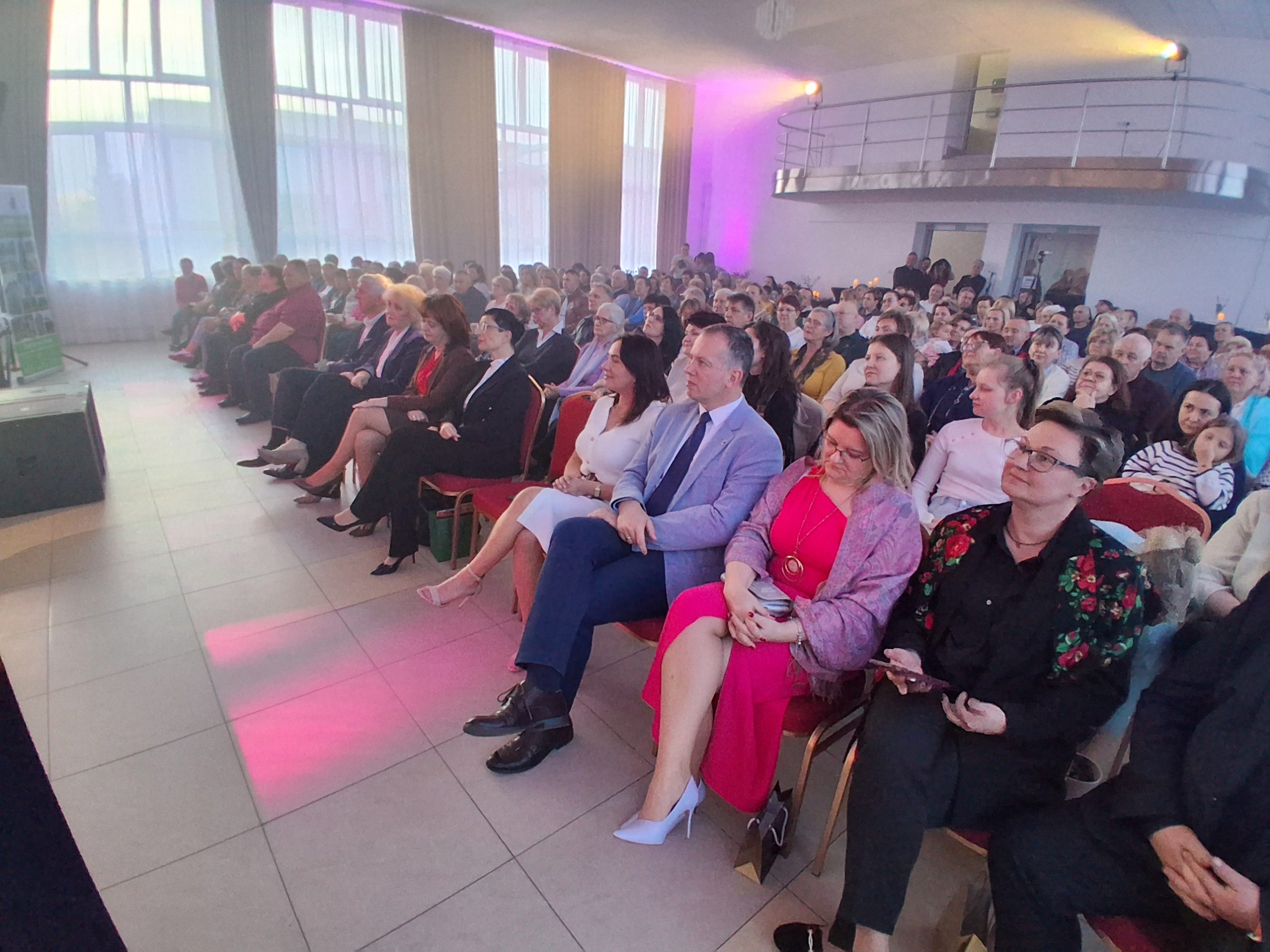 Na zdjęciu widać grupę ludzi siedzących w sali konferencyjnej. Są ubrani elegancko; dominują czerwone i białe akcenty. W tle duże okna i fioletowe oświetlenie.