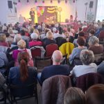 20260315_172042 Sala pełna ludzi siedzących na krzesłach, zwróconych w stronę sceny. Na scenie mężczyzna przemawia, dekoracje w jasnych kolorach. W tle oświetlenie w fioletowych odcieniach.