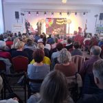 20260315_172215 Na zdjęciu widoczna jest sala pełna ludzi zwróconych w stronę sceny. Scena ozdobiona kolorowymi światłami i dekoracjami. Na scenie widoczni są dwaj mówcy.