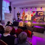 20260315_172652 Na scenie z fioletowym oświetleniem występuje kobieta i mężczyzna, dekoracje są kolorowe. W tle perkusja. Na widowni siedzą ludzie.