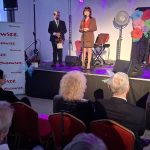 20260315_172703 Kobieta w brązowym kostiumie stoi na scenie z mikrofonem. Obok niej mężczyzna w garniturze, tło dekorowane kwiatami. Na widowni kilku widzów, po lewej baner z napisem Mazowsze.