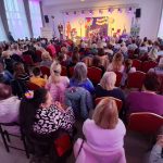 20260315_172732 Na zdjęciu jest pełna sala ludzi siedzących na czerwonych krzesłach. Na scenie znajdują się osoby, przy jasnym, kolorowym oświetleniu. Tło zdobią wiszące dekoracje, a za oknami widać zasłony.