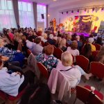 20260315_172802 Sala pełna ludzi siedzących na czerwonych krzesłach, scena oświetlona fioletowym światłem. W tle zasłony i stoliki. Na scenie stoi osoba, obok zestaw perkusyjny.