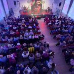 20260315_173347 Sala z publicznością siedzącą na czerwonych krzesłach, patrzącą na scenę z zespołem muzycznym. Tło jest jasne, z dużymi oknami po bokach i kolorowymi światłami. Dekoracje na scenie w intensywnych kolorach, atmosfera koncertowa