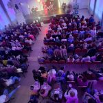 20260315_173606 Widok z góry na dużą salę z publicznością skierowaną w stronę sceny. Ludzie siedzą na czerwonych krzesłach w jasnym pomieszczeniu z kolorowymi światłami. Na scenie widać zespół muzyczny.