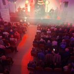 20260315_181227 Sala koncertowa w czerwonym oświetleniu. Widownia siedzi przed sceną z muzykami. Tło jest jasne z elementami dekoracyjnymi.